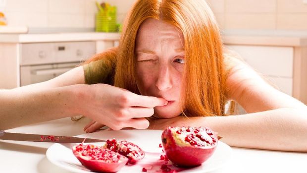 Anorexic young girl eating pomegranate with disgust in kitchen