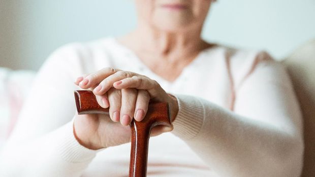 Elder holds hands on walking stick while sitting on white sofa in nursing home