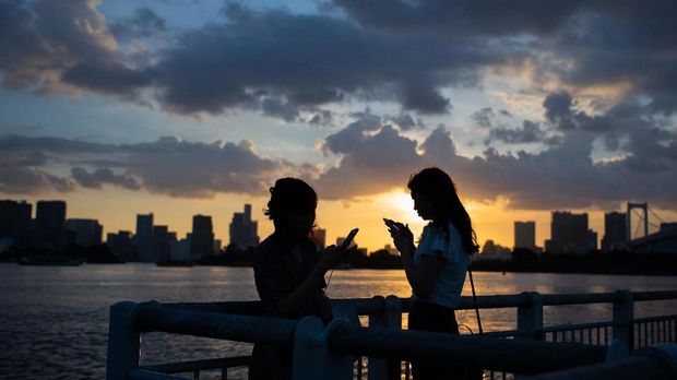 Women look at their phones during sunset in Odaiba, Tokyo on July 29, 2018. / AFP PHOTO / Martin BUREAU