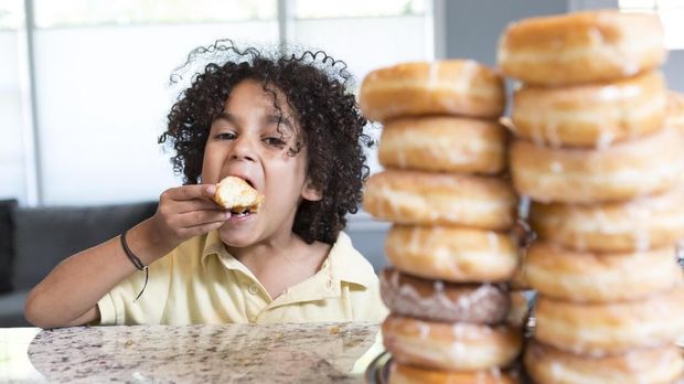 A little boy eats a donut and looks at the camera. He has a huge pile of donuts in front of him. He is hispanic and has big natural hair. He is sitting at a kitchen counter inside a home.