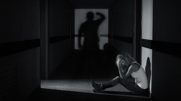 Young woman sitting in empty hallway covering her eyes under the shadow of a man