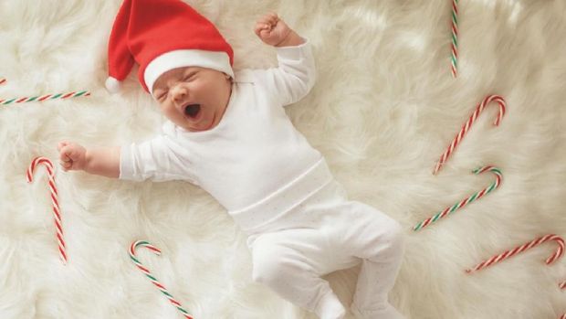 Cute little baby boy in Santa hat sitting inside of the red gift box next to the Christmas tree.