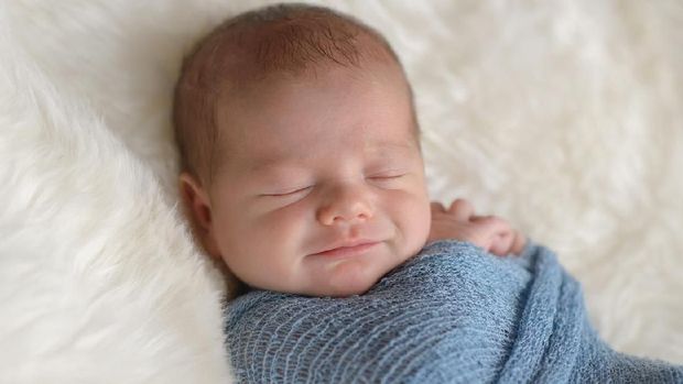 Sleeping, nine day old, newborn baby boy with a smile on his face. He is swaddled in a light blue wrap. Shot in the studio on a white, sheepskin rug.