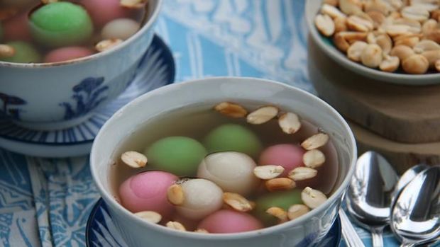 A Chinese Peranakan dessert of glutinous rice balls in warm ginger soup. Served in vintage ceramic bowls with extra peanuts, also in vintage saucer. A few teaspoons are placed on a folded batik napkin. The meal is arranged on a rustic wooden table.