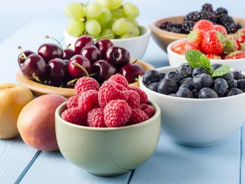 A selection of different Summer fruits, in a variety of bowls on a painted blue wood planked farmhouse kitchen table, against a light blue background.