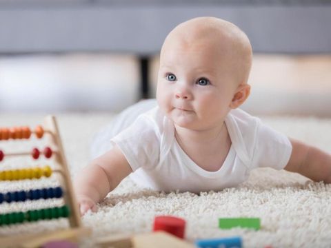 An adorable baby lays on her tummy on a rug in a living room.  She looks up with pursed lips.  There are wooden toys in the foreground.