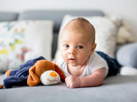 An adorable baby lays on her tummy on a rug in a living room.  She looks up with pursed lips.  There are wooden toys in the foreground.