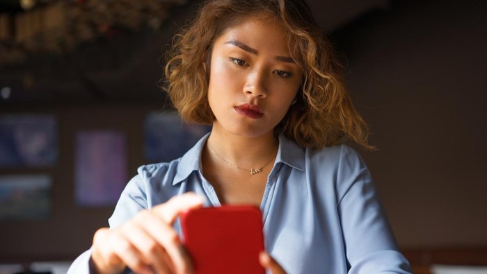Closeup of thoughtful young Asian woman holding mobile phone and surfing Internet. Attractive student taking selfie at cafe. Communication and work balance concept