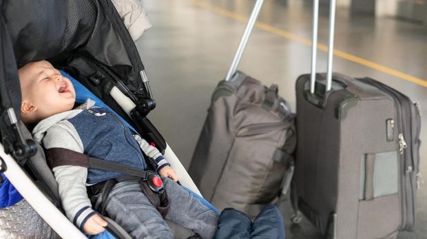 Screaming baby boy sitting in stroller near luggage at airport terminal. Child in carriage near check-in desk counter. Children tears, panic and hysterics. Travelling with small children concept.