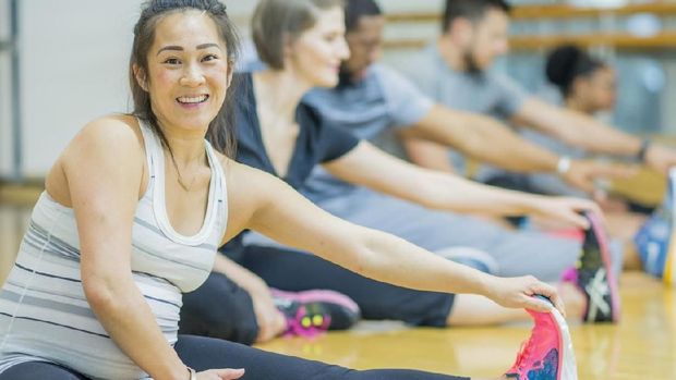A multi-ethnic group of adult men and women are indoors in a gym. They are wearing casual exercise clothing and running shoes. They are doing stretches by touching their toes. A pregnant Asian woman is smiling at the camera.
