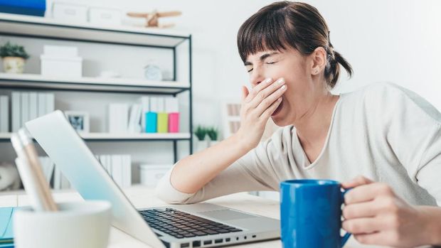 Tired sleepy woman yawning, working at office desk and holding a cup of coffee, overwork and sleep deprivation concept