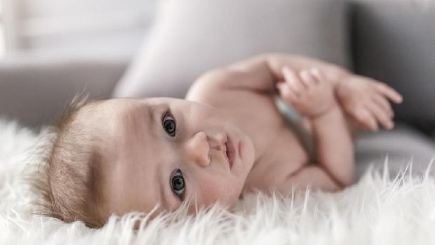 Close up portrait of infant lying on white fluffy bed indoors. Photo of the boy at the age of four months. Baby boy wearing diaper in white sunny bedroom. Newborn child relaxing in bed. Nursery for children. Textile and bedding for kids. Family morning at home. New born kid during side time looking at the camera.