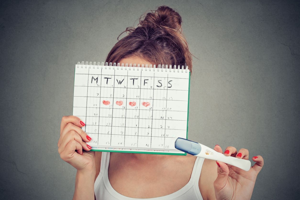 Young woman hiding behind a periods calendar and showing a positive pregnancy test