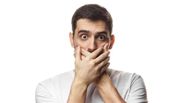 Indoor portrait of young European Caucasian man pictured isolated on white background dressed in white T-shirt, covering mouth with both palms, his brown eyes round with shock or deep astonishment