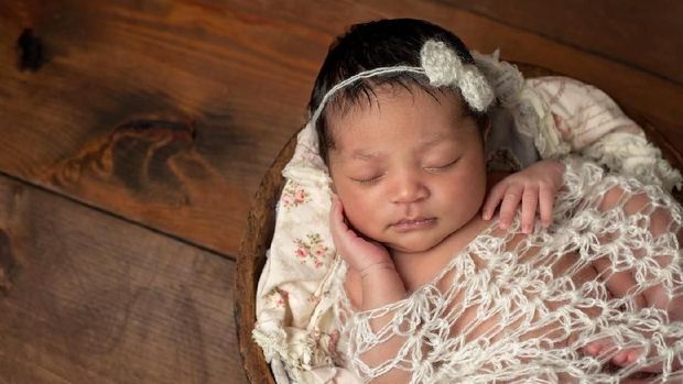A three week old newborn baby girl sleeping in a little, wooden bowl. She is wearing a cream colored bow headband and swaddled with a decorative wrap. Shot in the studio on a wood background.
