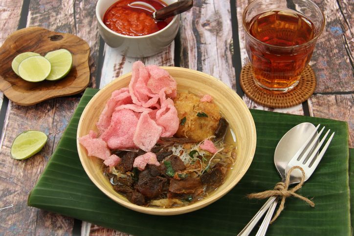 Soto Padang, the traditional beef jerky soup from Padang, West Sumatra. Crispy beef jerky with potato patty and glass noodles served in clear, spicy beef broth. Accompanied with pink tapioca crackers. The soup is plated in a ceramic bowl and placed on a plate lined with banana leaf. A set of rustic cutlery is placed on the plate.