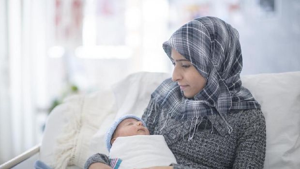 A muslim woman holds her newborn baby in her arms while sitting in a hospital bed. She is smiling at her son and holding him with such care.