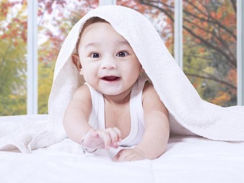 Portrait of joyful male infant crawling on the bed while smiling at the camera under a towel