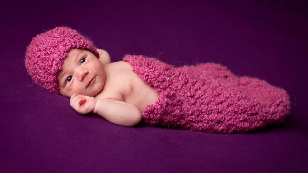 Color image of a precious, awake newborn baby girl, wearing a crocheted had and wrapped in a blanket, with purple background.