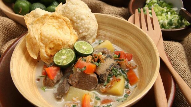 Soto Betawi, the popular traditional beef and offal soup from Betawi, Jakarta. The soup dish has been pre-plated in a bamboo bowl with an earthenware plate as the underliner. A pair of wooden spoon and fork is placed on the plate. The soup is surrounded with some of the condiments.