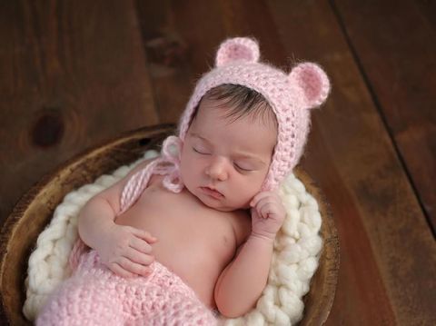 An overhead shot of a two week old newborn baby girl sleeping in a little, wooden bowl. She is wearing a crocheted, pink bear bonnet. Shot in the studio on a wood background.