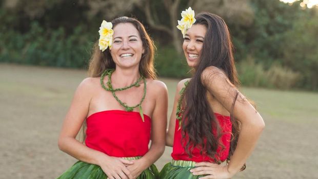 Two beautiful young local women practice hula on a beautiful day outside. They're standing together and laughing together at something happening off-camera.