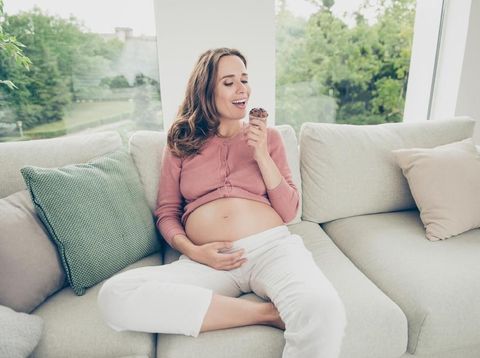Lovely nice beautiful adorable funny cheerful positive pregnant curly-haired mum in casual wear sitting on sofa, couch, divan, eating yummy cake, white light interior room