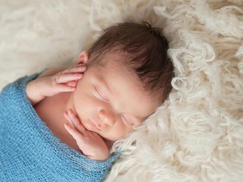 A 12 day old newborn baby boy sleeping on a beige flokati rug and swaddled in a gauzy light blue wrap.