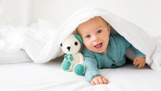 Little cute baby boy, child in knitted sweater, holding knitted toy, smiling happily at camera in white sunny, bright bedroom