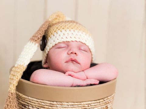 Front view of a cute white Caucasian newborn baby boy sleeping in a decorative basket, hands under the chin holding head, wearing a hand knitted long wool hat. Shot on Canon EOS into a studio. Warm toned edit to emphasize overall softness feel.