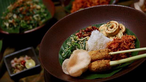 Nasi Campur Bali, the Balinese popular rice dish. Steamed white rice served with side dishes such as Sate Lilit (minced seafood satay), Ayam Pelalah (shredded chicken in red chili paste), Telur Dadar (egg omelet), Kacang Teri (peanuts and anchovies), Jukut Urap (vegetable salad with grated coconut dressing) and Kerupuk Udang (prawn crackers). The dish is served in a ceramic plate lined with banana leaf. The rice dish plate is in the focus with the buffet table being the blurry background.