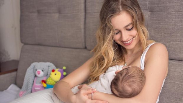 Breastfeeding baby. Pretty mother holding her newborn child. Mom smile and nursing infant. Beautiful woman and new born love at home. Blond mother breast feeding baby.