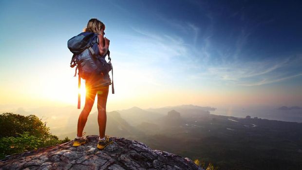 Young lady hiker standing with backpack on top of a mountain and enjoying sunrise
