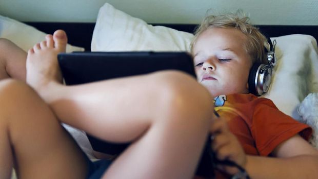 Boy wears headphones connected to a digital tablet while lying in bed. He looks at the content on the screen. It is night and he looks happy.  He could be streaming a movie, browsing the internet or playing a game.