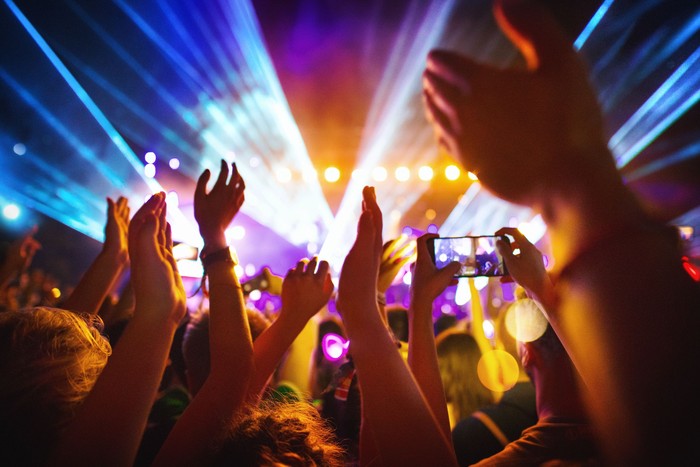 Rear view of excited crowd enjoying a DJ performance at a festival. There are many raised hands, some of the holding cell phones and taping the show.People in foreground are released.