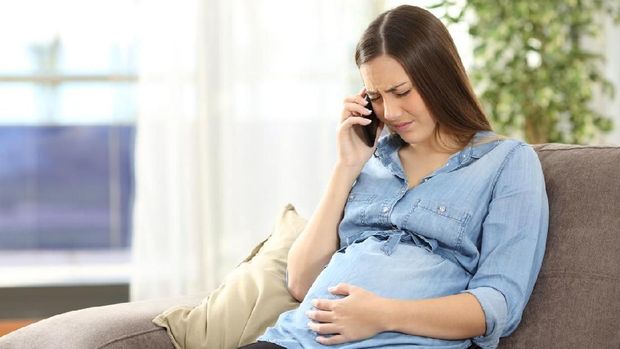 Young pregnant woman suffering belly ache and calling on the phone sitting on a couch in the living room in a house interior