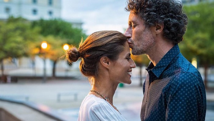 arti ciuman Mature husband kissing wife on forehead in the street in the evening. Romantic senior man giving a kiss to her woman in the city street. Loving middle aged couple in love at dusk.