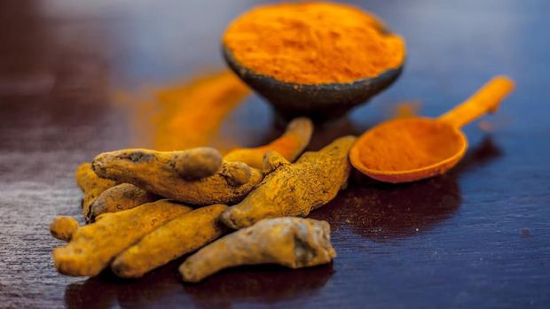 Raw organic famous and essential spice i.e. Turmeric or haldi or Curcuma or Curcuma, saffron des Indes in a clay bowl along with its powder on brown colored wooden surface.