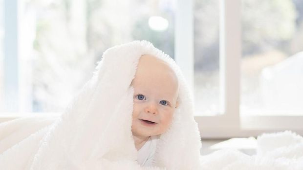 Beautiful 6 Month Baby Boy Dressed in White & Lying on Fluffy White Blanket Looking at Camera. Smiling & Happy