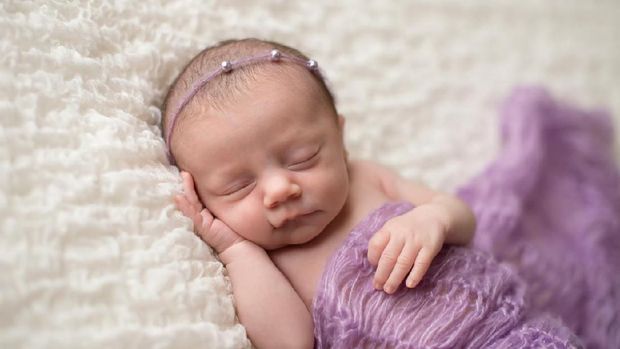 Sleeping, two week old newborn baby girl covered in a lavender purple blanket and wearing a pearl headband.