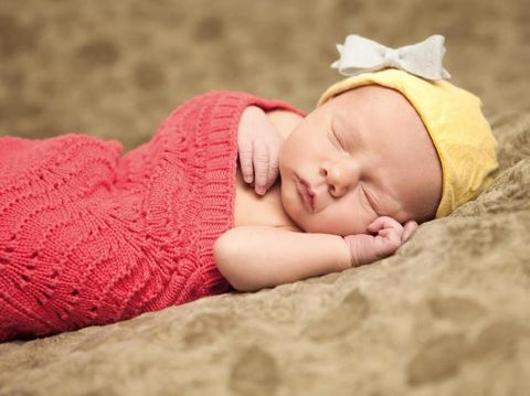 Portrait of a newborn baby with yellow hat and white bow, wrapped in red knit blanket.