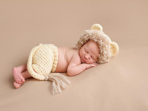 A smiling three week old newborn baby boy wearing a crocheted lion costume. Shot in the studio on a khaki colored background.
