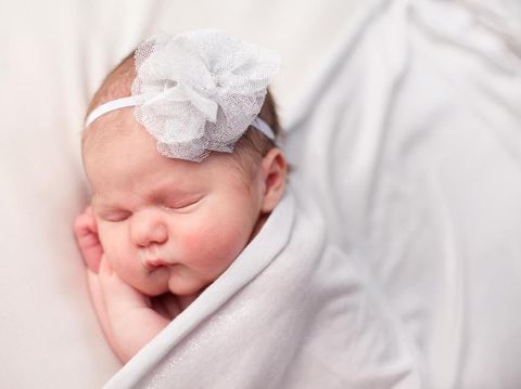 Color photo of a newborn baby girl wearing a headband and  sleeping peacefully, covered with a white blanket.