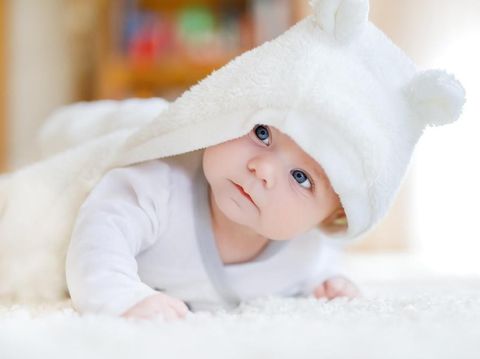 Baby girl with blue eyes wearing white towel or winter overal in white sunny bedroom. Newborn child relaxing in bed. Nursery for children. Textile and bedding for kids. New born kid with toy bear.