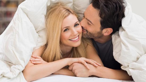 Handsome man kissing woman on cheek under a duvet in the bed. Happy young couple lying down comfortably in their bed at home. Young couple having fun lying on bed at home.