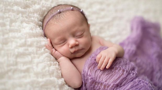 Sleeping, two week old newborn baby girl covered in a lavender purple blanket and wearing a pearl headband.