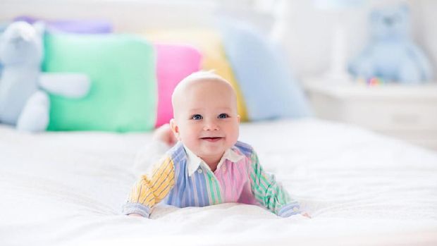 Baby boy in white bedroom. Newborn child in bed with pastel color cushions. Nursery for children. Textile, pillows and bedding for kids. Family morning at home. New born kid tummy time with toy bear.