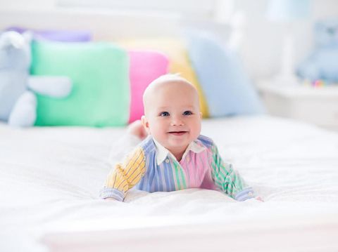 Baby boy in white bedroom. Newborn child in bed with pastel color cushions. Nursery for children. Textile, pillows and bedding for kids. Family morning at home. New born kid tummy time with toy bear.