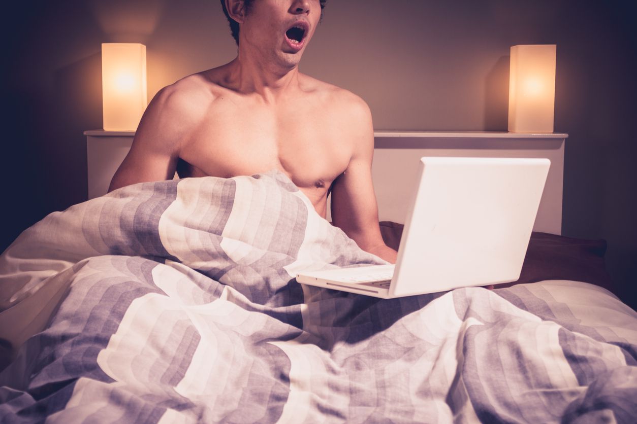 Young man is sitting in bed and watching pornography on his laptop