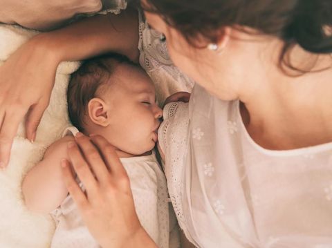 Young beautiful woman is breastfeeding a little baby in a cozy room. On the table there is a white Cup and a green plant. On the wall hang a white empty picture frames. No images of nudity